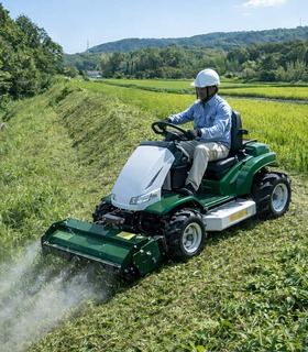 ヘルメットを着用した作業員が乗用草刈機（ハンマーナイフモア）を運転し、広大な圃場の緑肥や雑草を粉砕・刈り取る様子